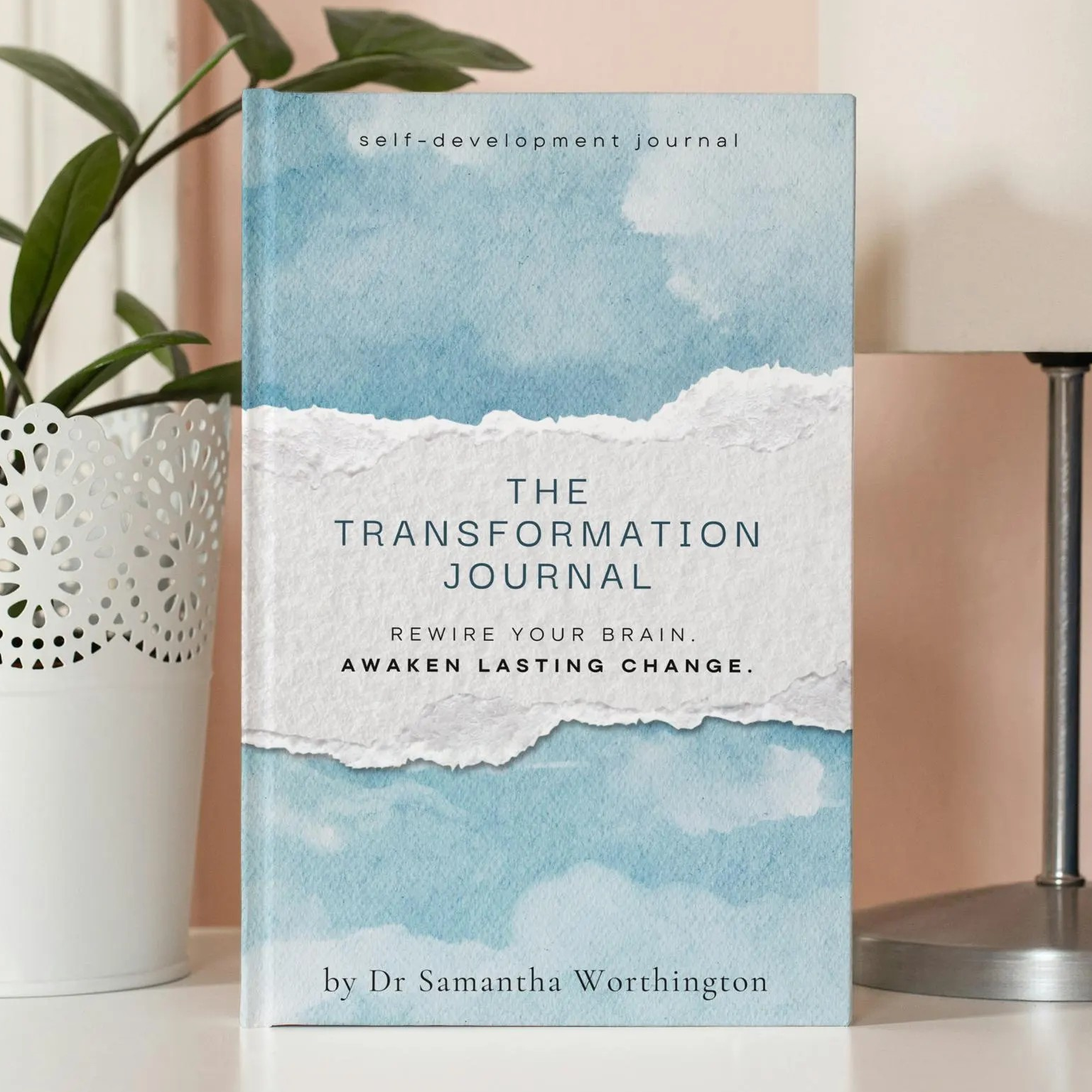 Book titled 'The Transformation Journal' on a surface with a lamp and plant in the background. Dr Samantha Worthington